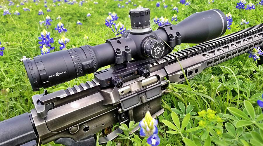 A close-up of a rifle with a mounted scope lying in a field of green grass and blue wildflowers.
