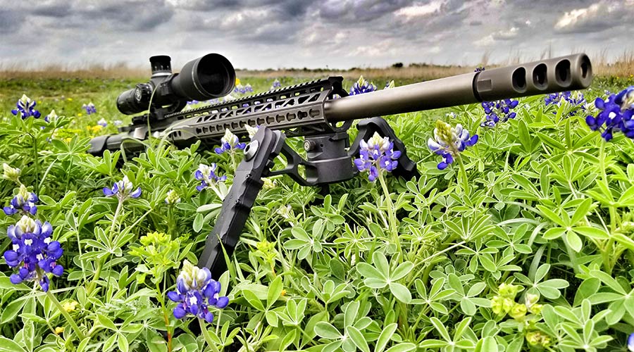 A scoped rifle with a bipod is positioned among green plants and purple flowers in a field, under a cloudy sky.