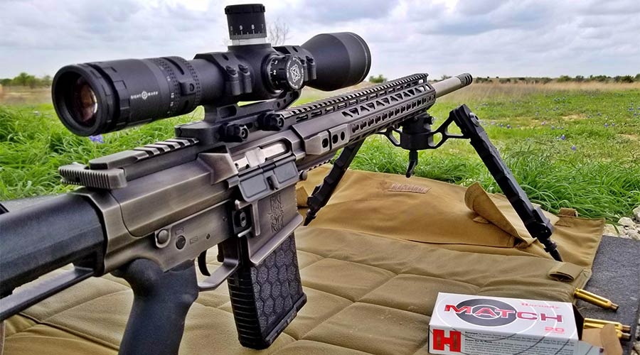 A scoped semi-automatic rifle with a bipod is set up outdoors on a tan shooting mat. A box of Hornady Match ammunition is visible next to the rifle, and grassy fields stretch into the distance.
