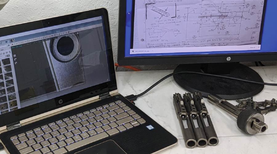 A laptop and monitor display technical diagrams. Metal tools and parts are arranged on a white desk nearby.