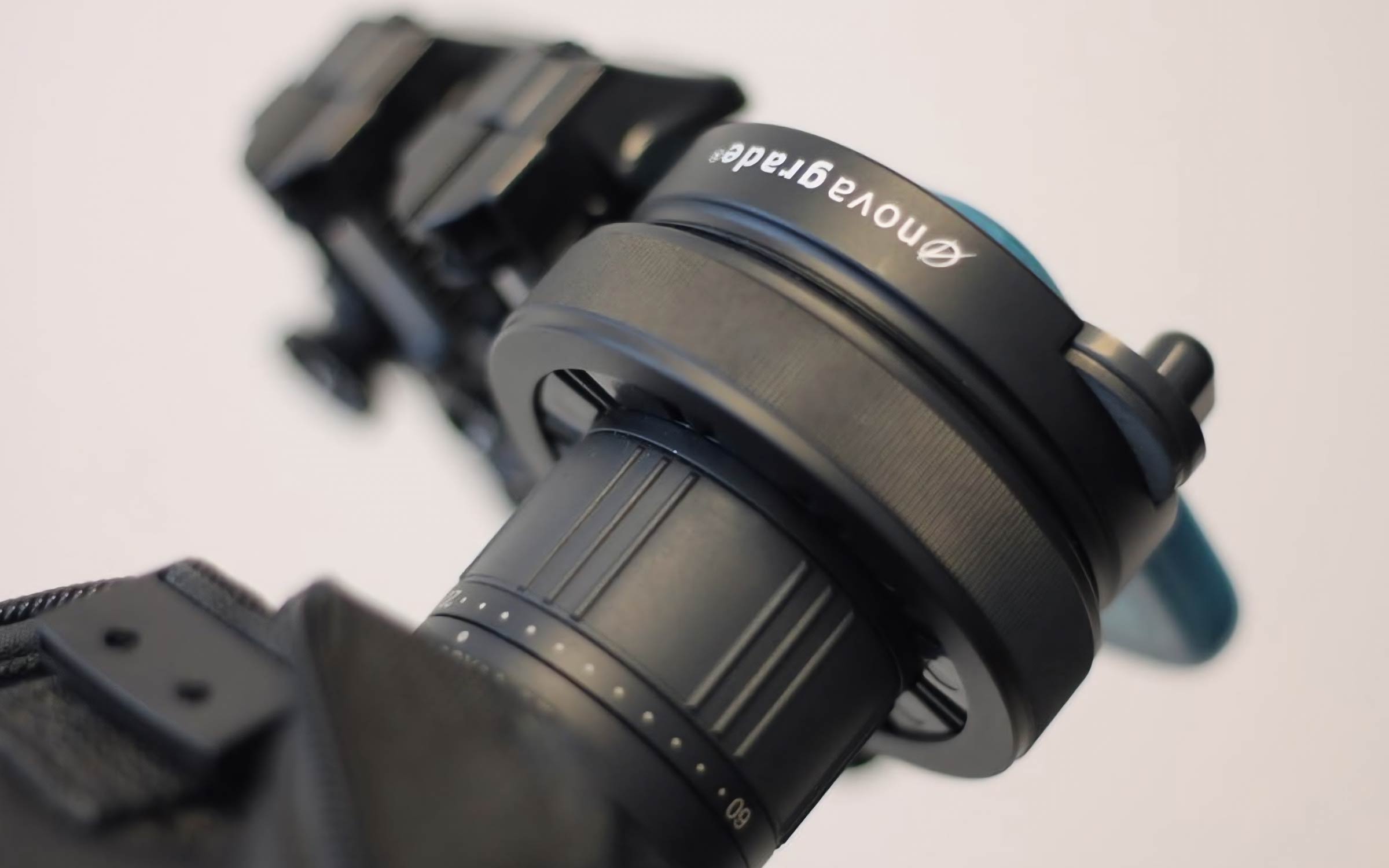 A close-up of a black camera lens with a blue lever and the word Novagrade printed on the lens adapter, against a plain background.