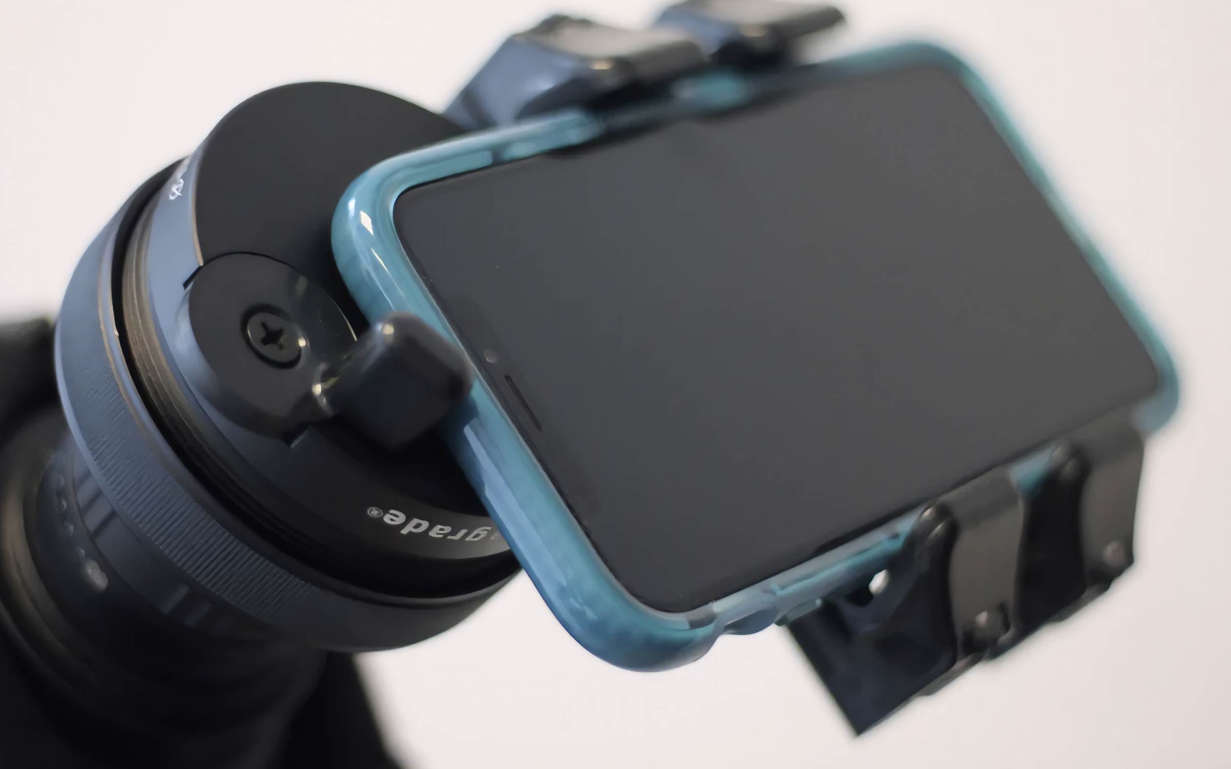 A smartphone in a blue case is mounted onto a camera lens using a clamp adapter, with a blurred background.