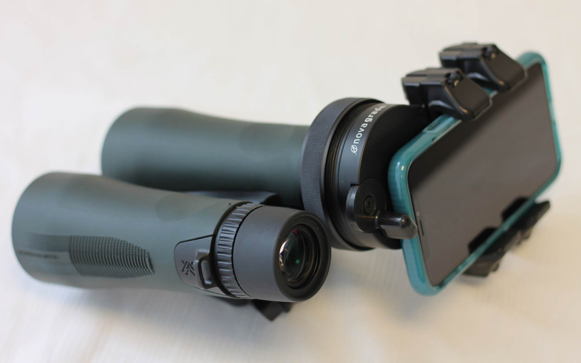 Two green binoculars are attached to a smartphone using a special mounting adapter, allowing the phone camera to take photos or videos through the binocular lens. All items are on a white surface.