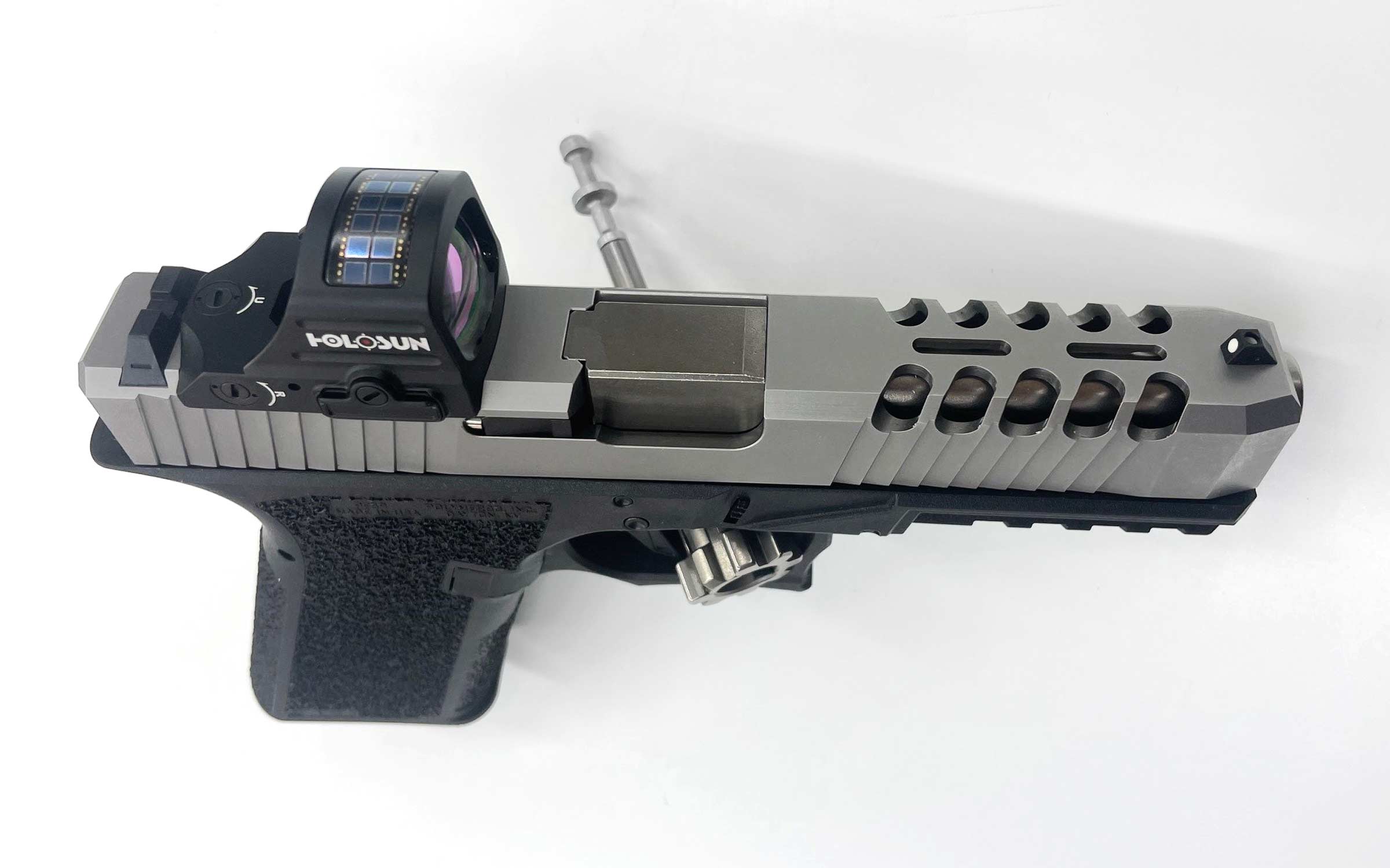A silver and black handgun with a textured grip, slide cuts, a mounted red dot sight, and a takedown tool inserted into the barrel, placed on a white surface.